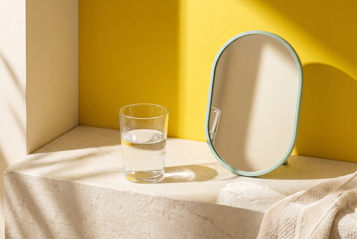 A glass of water and oval mirror on a sunlit stone ledge against a yellow wall.