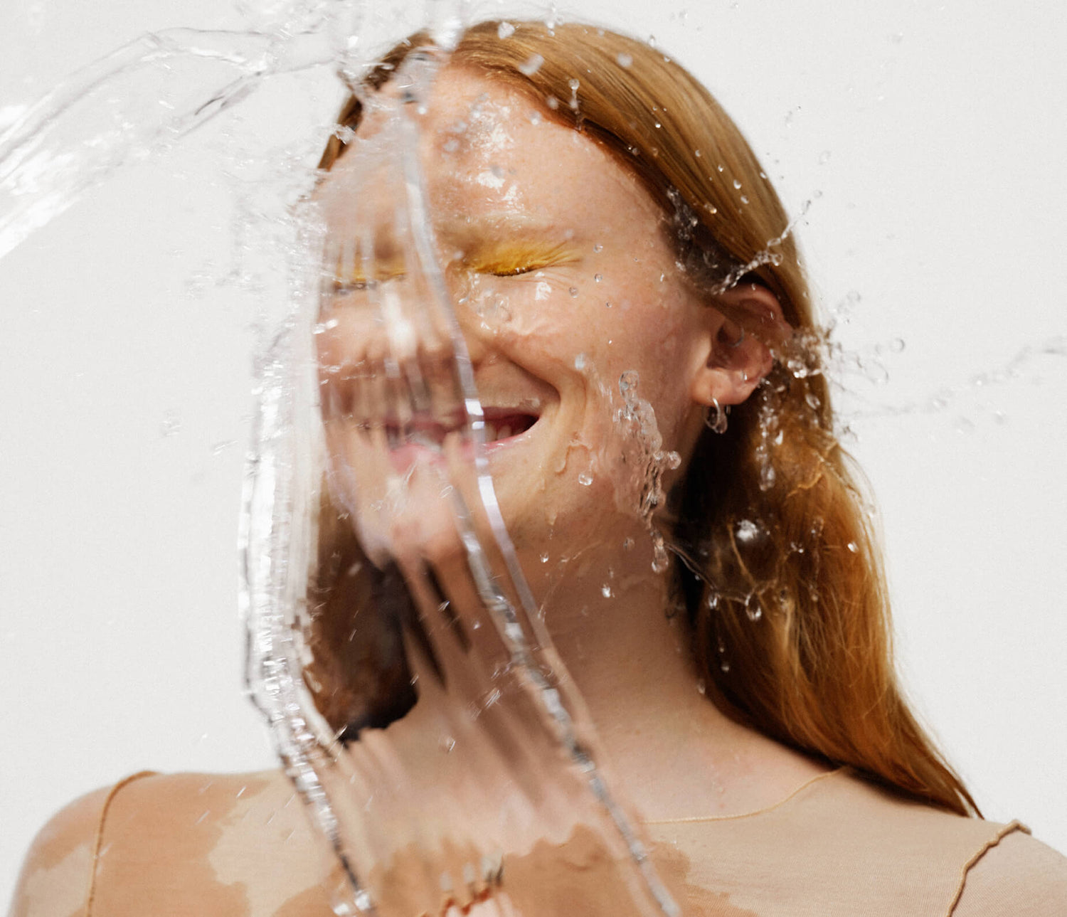 Woman with red hair and yellow eyeshadow smiling and splashing water on her face.