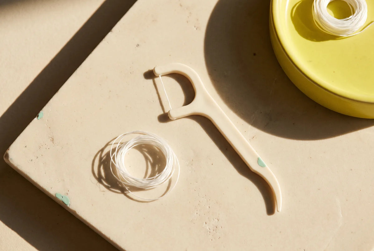 An ivory floss pick and loose white string floss on sunlit cream stone beside a yellow dish.